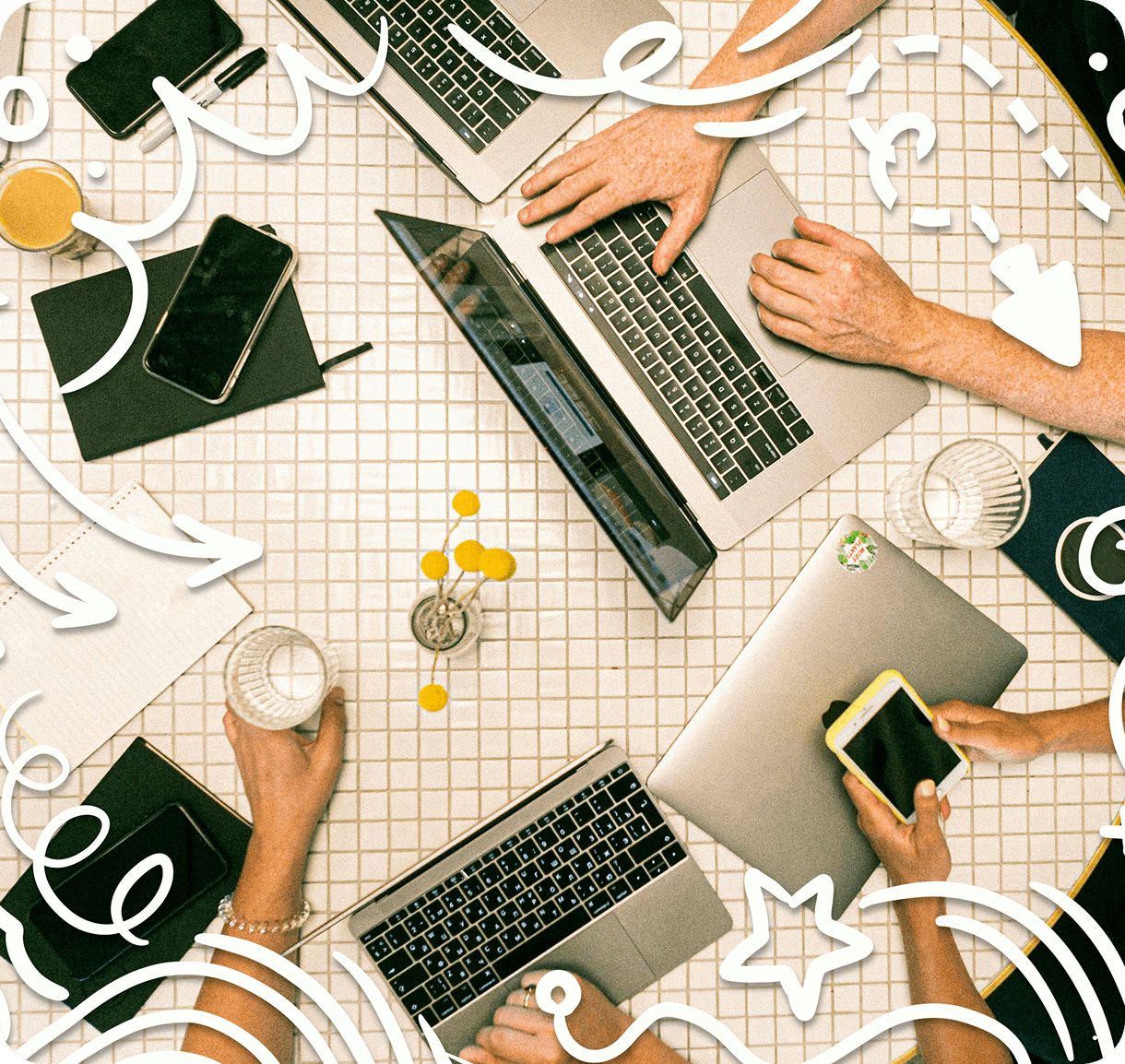 A top view of a table with laptops, smartphones, and hands interacting. The table has a grid pattern, and there are decorative yellow flowers. Artistic white doodles are overlaid, featuring arrows and swirls. Various office items and beverages are present.
